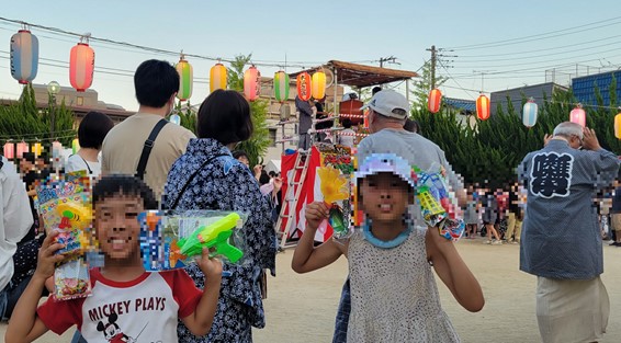 射的と100円くじのおもちゃを持った子供たちの後ろを盆踊りを踊る人たちが